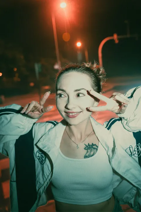 Kobieta w białej sportowej kurtce kuca na chodniku pod czerwoną latarnią podczas nocnej sesji miejskiej.